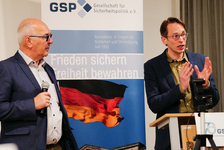 Zwei Männer bei einer GSP-Veranstaltung. Einer spricht am Rednerpult, der andere hält ein Mikrofon. Deutscher Flaggenhintergrund.