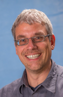 Porträt eines lächelnden Mannes mit kurzen grauen Haaren, Brille und grauem Hemd vor blauem Hintergrund.