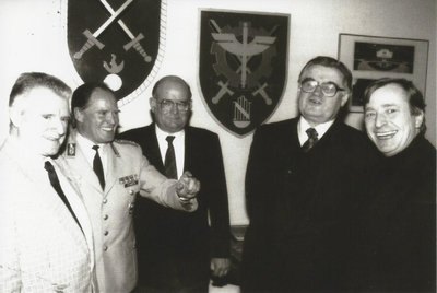 Fünf lächelnde Männer, einer in Militäruniform, vor Wandemblemen. Schwarz-Weiß-Aufnahme.