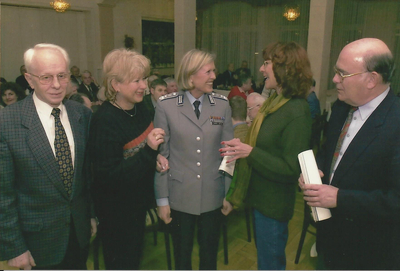 Fünf Personen, darunter eine Frau in grauer Militäruniform, lächeln und interagieren bei einer gesellschaftlichen Veranstaltung.