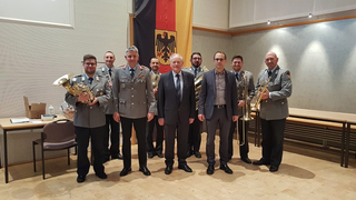 Gruppe Männer, einige in Uniform mit Blechblasinstrumenten, andere in Anzügen, vor deutscher Flagge stehend.