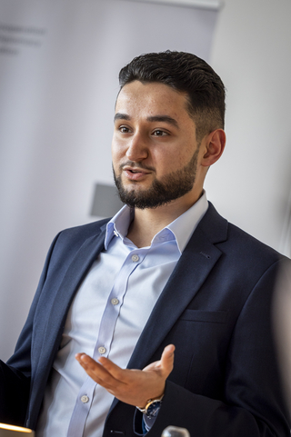 Junger bärtiger Mann in blauem Anzug und Hemd, gestikuliert sprechend vor hellem Hintergrund.