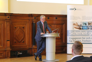 Ein Mann im Anzug spricht an einem Rednerpult vor einer Holzwand und einem Banner zur Sicherheitspolitik.