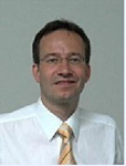 Porträt eines lächelnden Mannes mit Brille, kurzen dunklen Haaren, weißem Hemd und gelb gestreifter Krawatte.