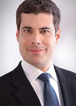 Kopfportrait eines lächelnden Mannes im dunklen Anzug, weißen Hemd und blauen Krawatte, blickt direkt in die Kamera.