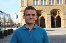 Ein junger Mann in einem blauen Poloshirt steht leicht lächelnd vor einem historischen Steingebäude mit Bögen.