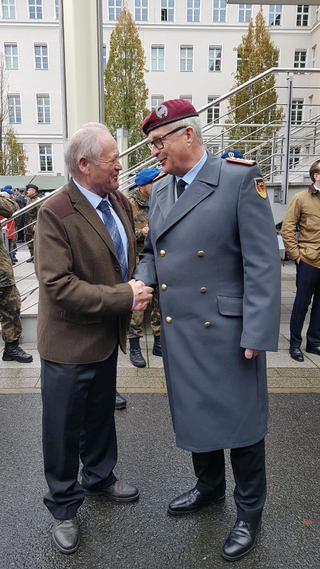 Zwei lächelnde Männer schütteln Hände, einer in Zivil, der andere in Bundeswehruniform mit rotem Barett.