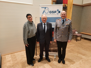 Drei Personen: Frau, Mann im Anzug, Mann in Militäruniform, vor GSP-70-Jahre-Banner und deutscher Flagge.