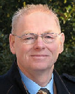 Kopfaufnahme eines lächelnden älteren Mannes mit kurzen hellen Haaren, Brille, blauem Hemd und gemusterter Krawatte.