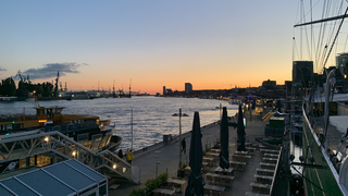 Erhöhte Ansicht eines Hafens bei Sonnenuntergang, mit breitem Fluss, festgemachten Schiffen, beleuchtetem Kai und Stadt im Hintergrund.