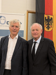 Zwei reife Männer in dunklen Anzügen posieren vor einer deutschen Flagge und einem sicherheitsbezogenen Banner.