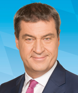 Porträt des deutschen Politikers Markus Söder, lächelnd, in dunklem Anzug mit rosa Krawatte vor blauem Hintergrund.