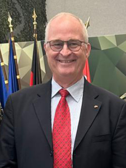 Ein lächelnder Mann mittleren Alters mit grauen Haaren und Brille, im dunklen Anzug mit roter Krawatte, vor Flaggen.