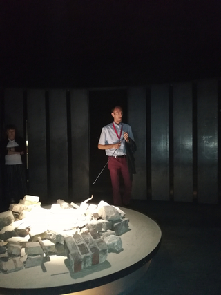 Museumsführer mit Zeigestab vor beleuchtetem Trümmer-Exponat in einem abgedunkelten Raum mit dunklen Wänden.