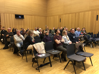 Vielfältiges Publikum von Erwachsenen in einem modernen Hörsaal mit Holzwänden, die einer Präsentation zusehen. Ein Projektor ist sichtbar.