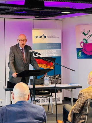 Ein Mann im Anzug spricht an einem Rednerpult vor einem GSP-Banner, mit Publikum im Vordergrund.