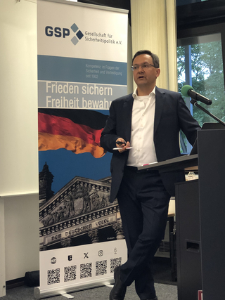 Ein Mann im Anzug spricht an einem Rednerpult vor einem Banner der Gesellschaft für Sicherheitspolitik mit deutscher Flagge und Reichstag.