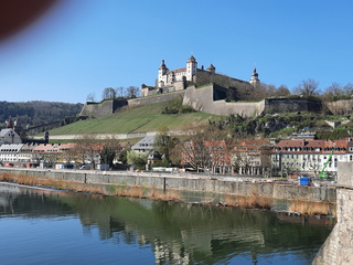 Festung Marienberg in Würzburg auf einem grünen Hügel mit Weinbergen, die Stadt und der Main im Vordergrund unter klarem blauem Himmel.