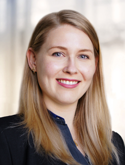 Professionelles Porträt einer lächelnden blonden Frau mit blauen Augen in einem schwarzen Blazer und blauen Hemd.