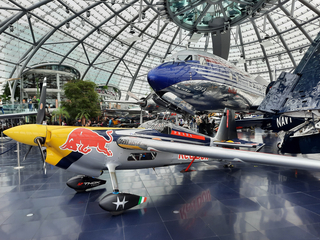 Ein Red Bull Kunstflugzeug ausgestellt in einem modernen Glashangar, mit anderen großen Flugzeugen wie DC-6 und Corsair im Hintergrund.