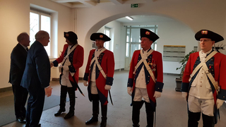 Zwei Männer in Anzügen schütteln die Hand eines Mannes in historischer Militäruniform, während vier weitere in Uniformen danebenstehen.