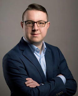 Porträt eines Mannes mit Brille, blauem Anzug und hellblauem Hemd, der mit verschränkten Armen in die Kamera blickt.