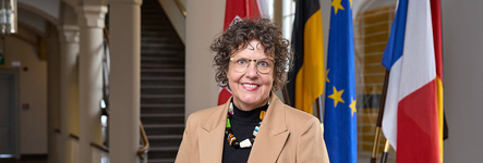 Lächelnde Frau mit lockigem Haar und Brille, beigem Blazer und bunter Kette, vor europäischen Flaggen.
