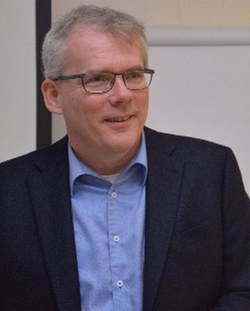 Ein Mann mit grauem Haar und Brille, der einen dunklen Anzug und ein blaues Hemd trägt, lächelt leicht.