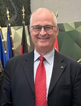 Ein lächelnder Mann mit Brille, dunklem Anzug und roter Krawatte steht vor Flaggen und einem geometrischen Hintergrund.