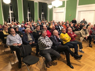 Ein großes Publikum aus Erwachsenen und Senioren sitzt in einem Saal mit grünen Wänden bei einer Veranstaltung.