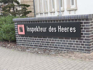 Schild mit der Aufschrift „Inspekteur des Heeres“ und einem Wappen, eingebettet in eine Ziegelmauer vor einem Gebäude.