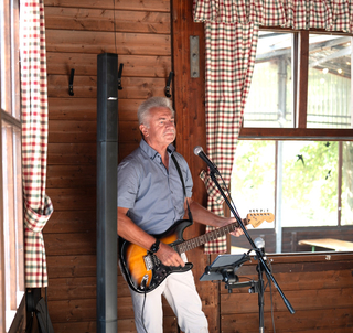 Älterer Mann mit grauen Haaren spielt E-Gitarre in rustikalem Raum mit Holzwänden, karierten Vorhängen und Mikrofon.