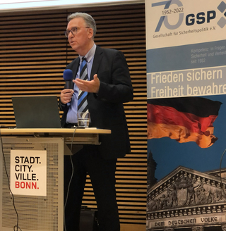Ein Mann spricht an einem Pult mit Mikrofon, Laptop und einem 'STADT. BONN.' Schild. Ein GSP-Banner ist hinter ihm.