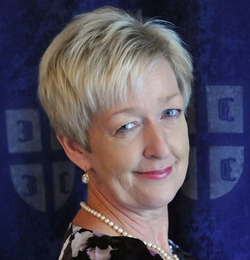 Porträt einer lächelnden Frau mit kurzen blonden Haaren, blauen Augen, einer Perlenkette und einem gemusterten Oberteil.