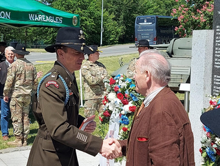 Militäroffizier in Uniform und Zivilist schütteln Hände bei einer Gedenkfeier mit Kranz und Soldaten im Hintergrund.