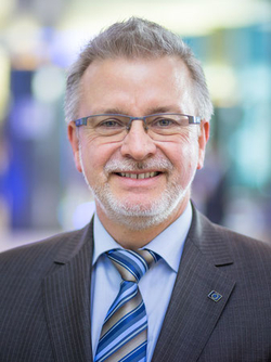 Porträt eines lächelnden Mannes mittleren Alters mit grauem Haar, Bart, Brille und einem Nadelstreifenanzug mit gestreifter Krawatte.