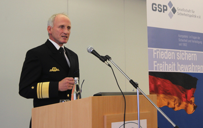 Ein Marineoffizier spricht am Pult, mit deutscher Flagge und 'GSP Gesellschaft für Sicherheitspolitik' Banner.