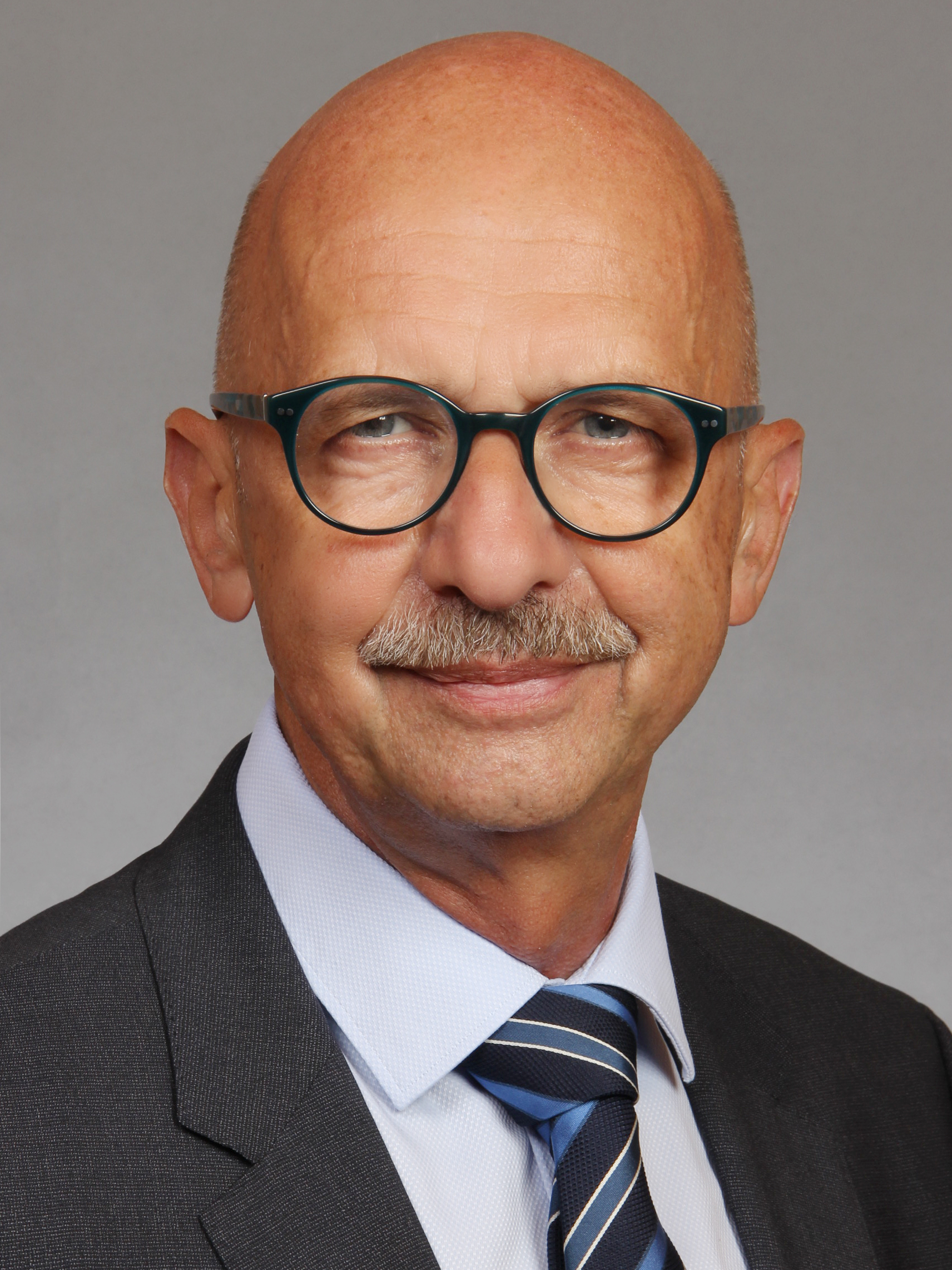 Porträt eines lächelnden, glatzköpfigen Mannes mittleren Alters mit Schnurrbart und tealfarbener Brille, im Anzug.