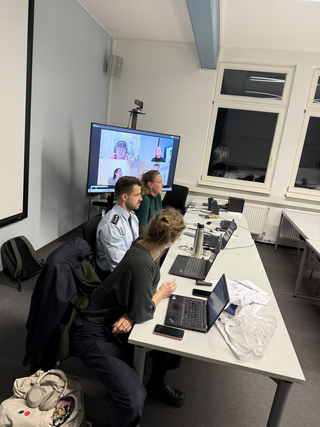 Drei Personen sitzen an einem Tisch in einem Raum und nehmen an einem Hybrid-Meeting mit Videokonferenz teil.