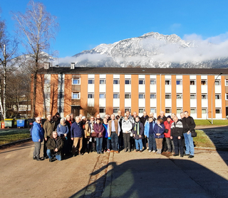 Eine Gruppe Erwachsener steht vor einem großen Gebäude mit schneebedeckten Bergen und blauem Himmel im Hintergrund.