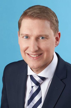 Lächelnender Mann mit hellbraunem Haar und blauen Augen, in dunkelblauem Anzug und gestreifter Krawatte vor blauem Hintergrund.