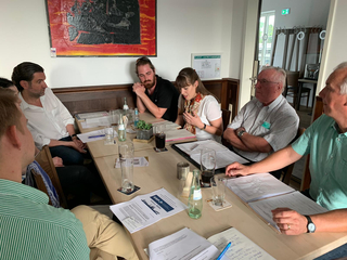 Vielfältige Gruppe bei einem informellen Treffen mit Dokumenten und Getränken, über ein Thema (vermutlich GSP station Köln) diskutierend.