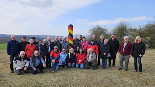Gruppe von etwa 30 Erwachsenen, meist Senioren, posiert im Freien vor einem deutschen Grenzstein auf einer Wiese.