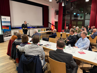Ein Marineoffizier spricht vor einem Publikum bei einer Veranstaltung der Gesellschaft für Sicherheitspolitik, mit Banner und Leinwand.