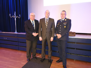 Drei Männer, zwei in Anzügen und einer in dunkler Uniform mit Medaillen, stehen auf einer Bühne mit blauem Vorhang und weißer Leinwand.