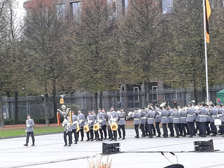 Eine Militärkapelle in Uniform paradieren auf einem gepflasterten Platz, mit Musikern und einer teilweise sichtbaren Deutschlandflagge.