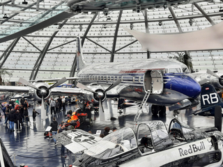 In einem modernen Hangar sind Red Bull Flugzeuge und ein Formel-1-Wagen ausgestellt, umgeben von Besuchern.
