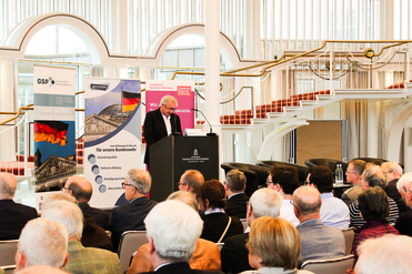 Jean Asselborn spricht an einem Rednerpult vor einem Publikum in einem hellen Konferenzsaal mit deutschen Flaggen und Bannern.