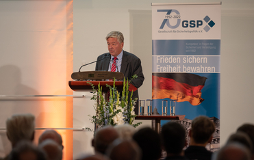 Mann am Rednerpult des Bayerischen Landtags spricht vor Publikum; GSP-Banner mit 'Frieden sichern Freiheit bewahren'.
