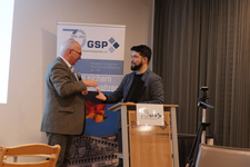 Zwei Männer, einer mit Mikrofon, reichen sich an einem Podium vor einem GSP-Banner die Hände.